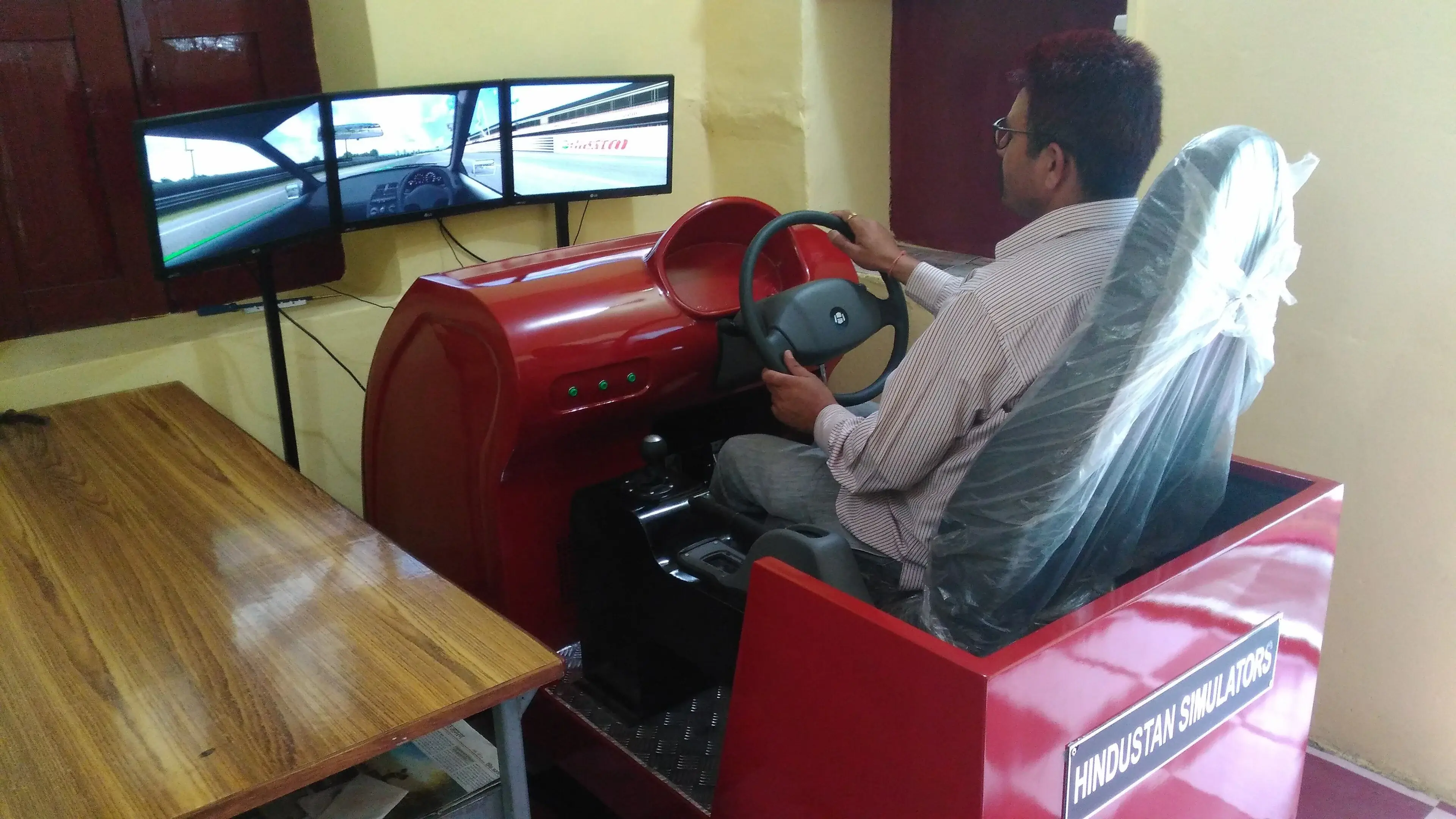 A seated driver holds a steering wheel in front of a three-screen driving simulator.