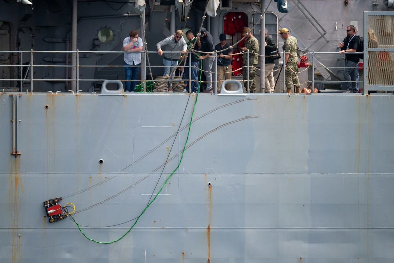 Gecko Robotics Toka 4 inspection crawler on the outside hull of a Navy test ship, with engineers and sailors watching from the deck.
