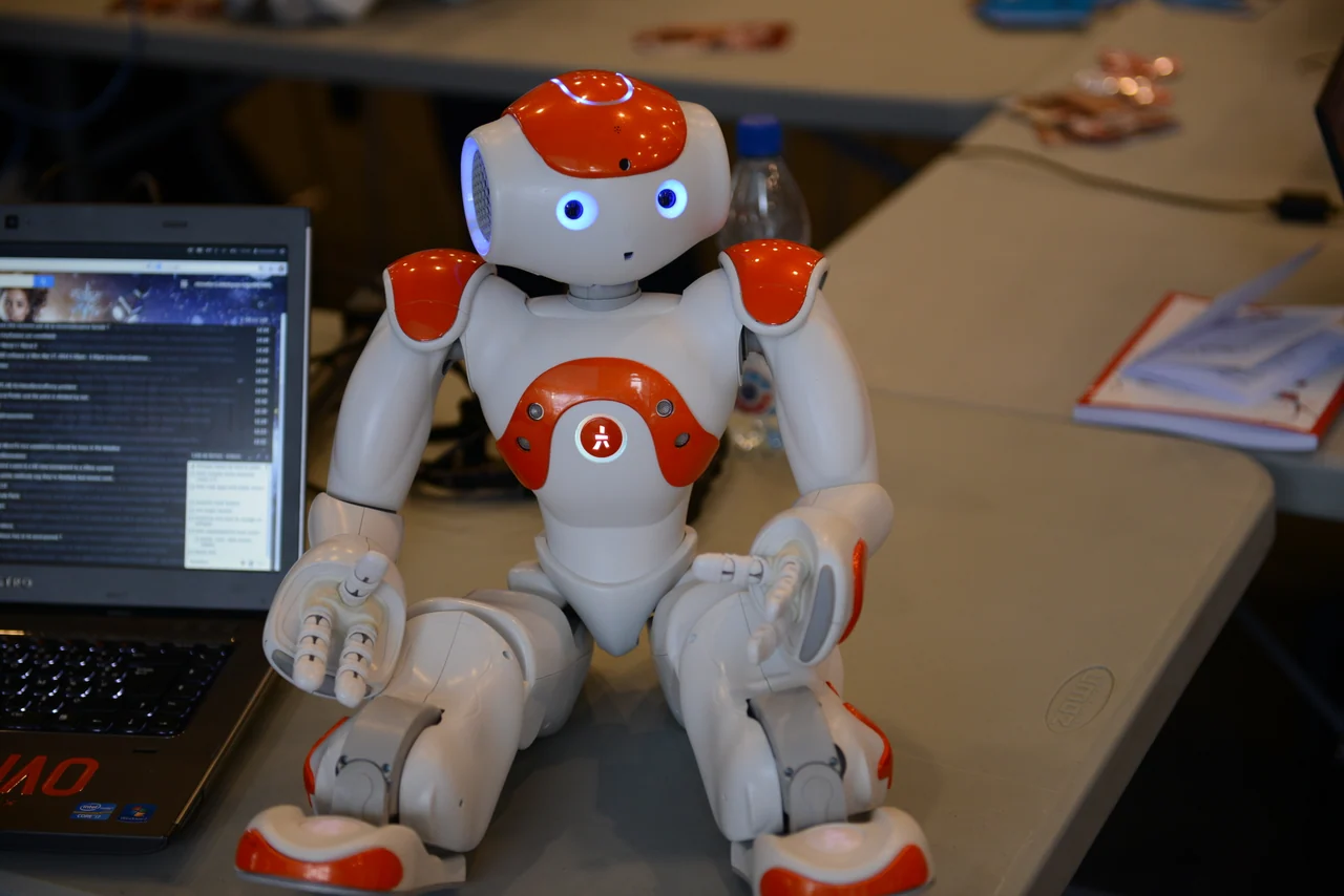 A friendly red and white NAO humanoid robot beside a laptop at a robotics workshop.